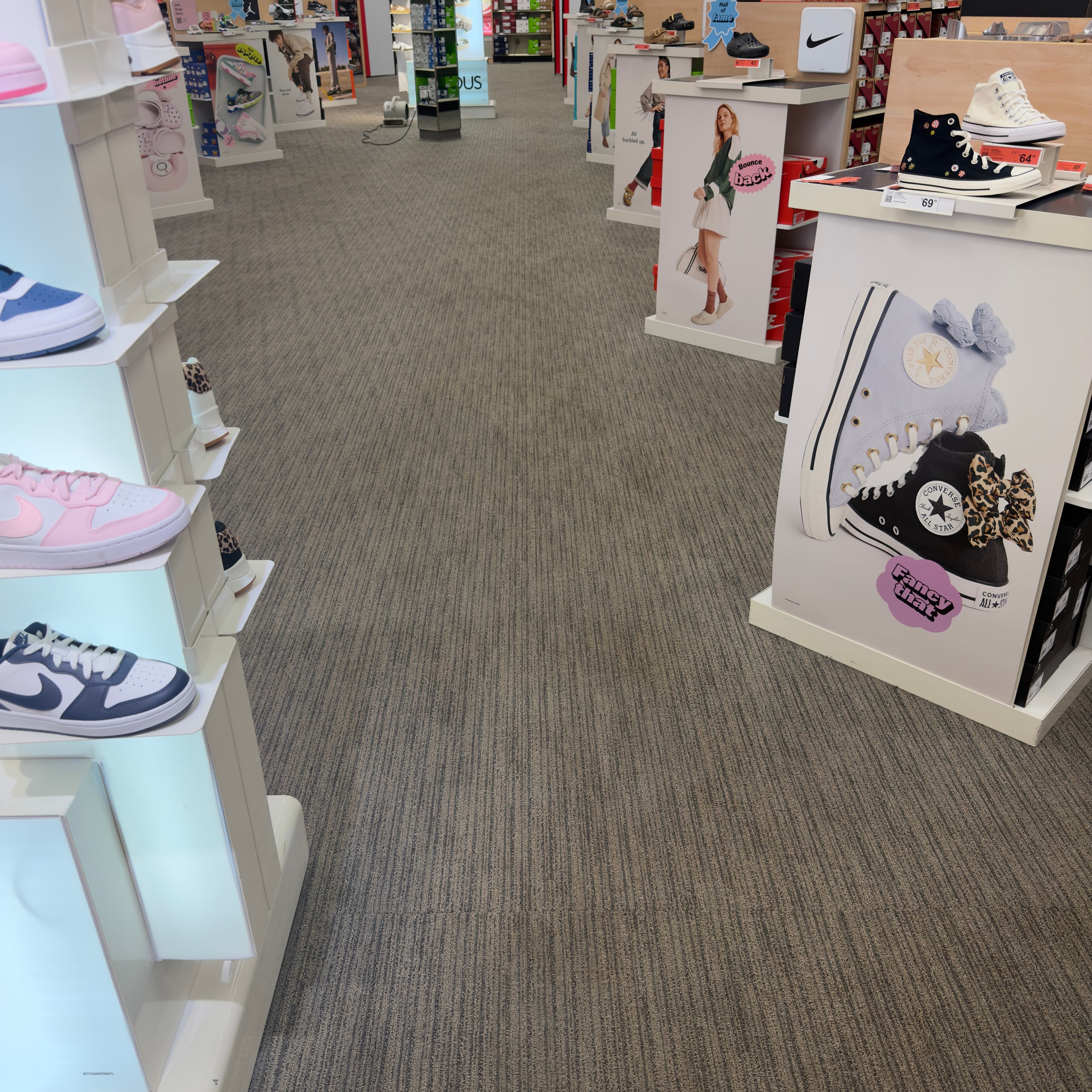 Shoe store aisle with sneaker displays featuring Nike and Converse shoes on grey textured carpet.