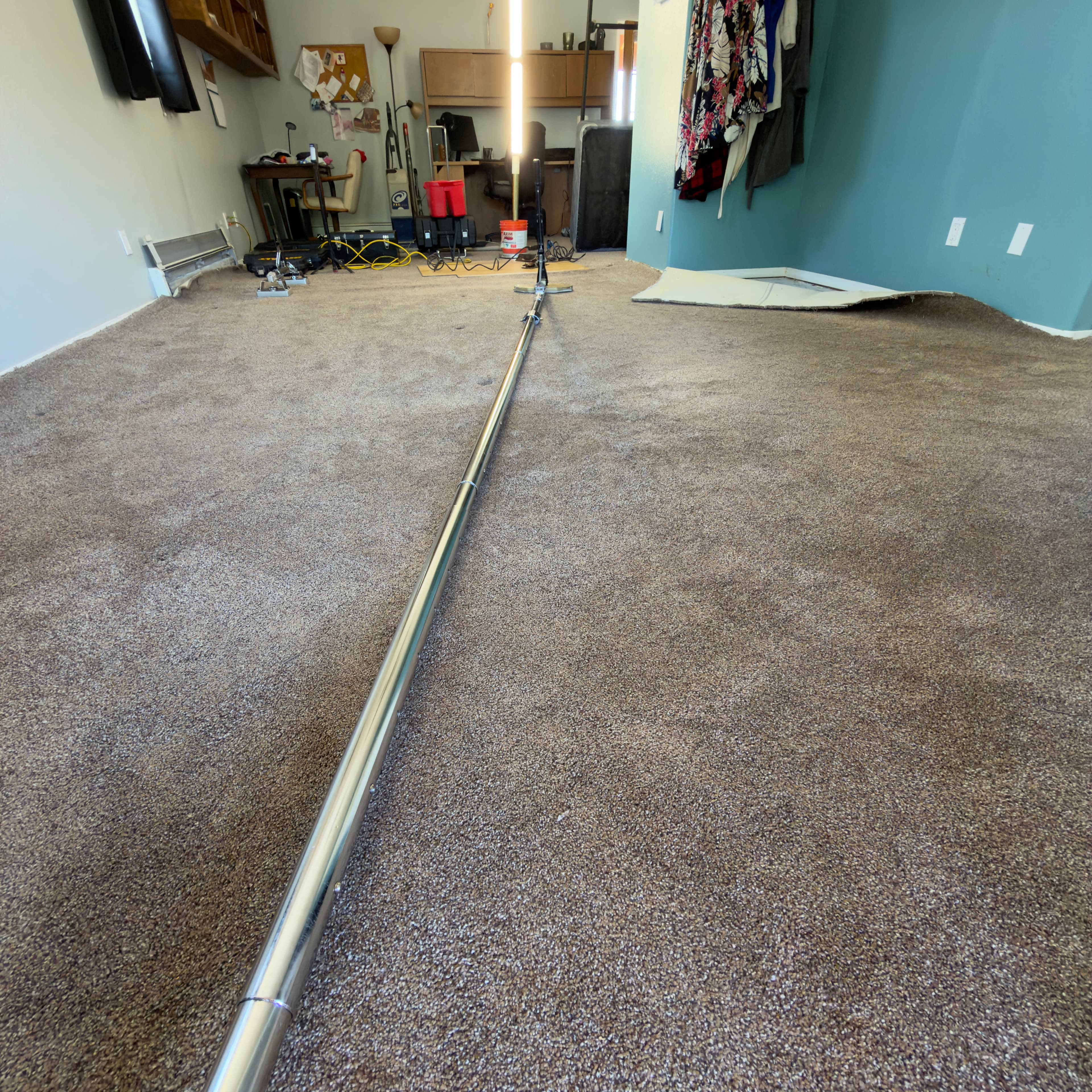Long silver carpet stretcher tool extending across a brown carpeted floor in a cluttered room.