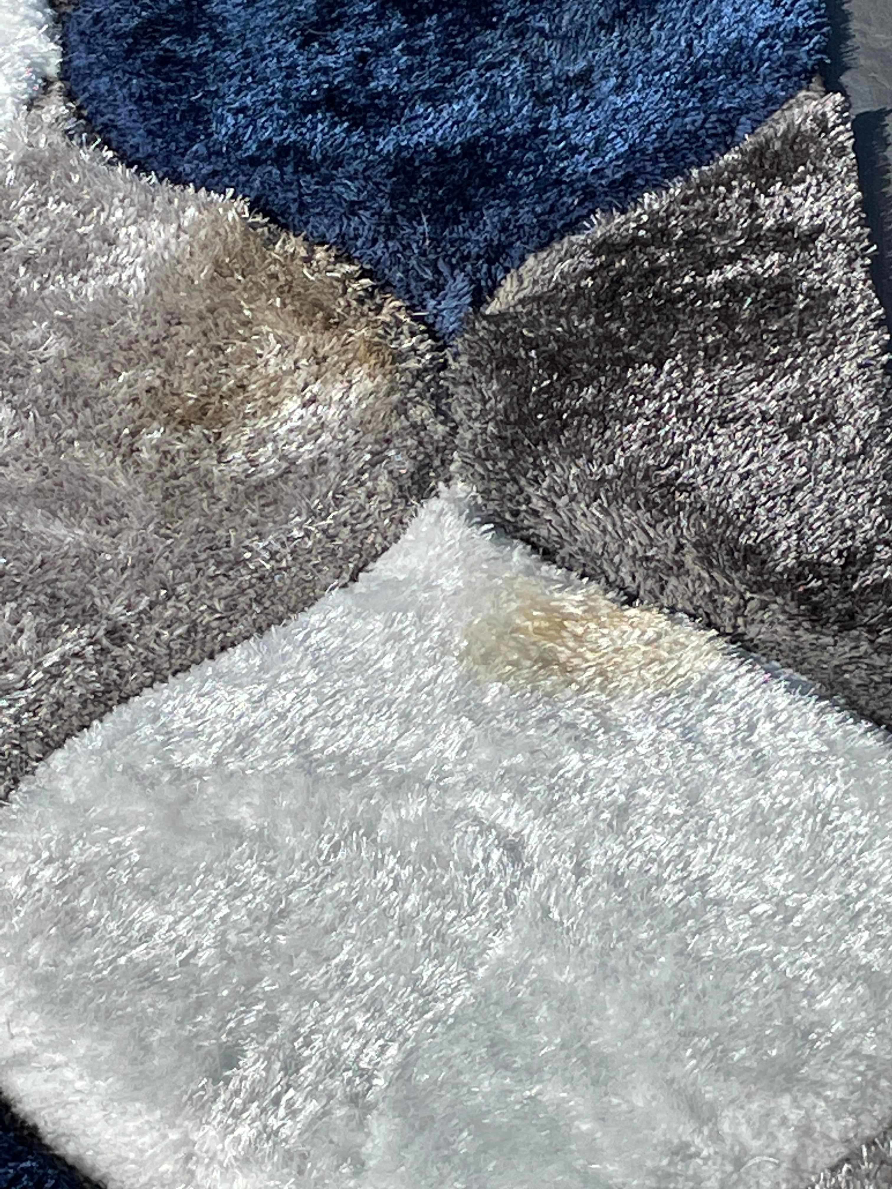 Close-up of a plush geometric rug featuring blue, gray, and white sections with a stain.