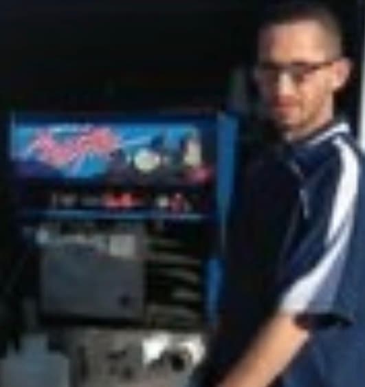 Man with glasses and a striped shirt standing next to a blue retro arcade cabinet.